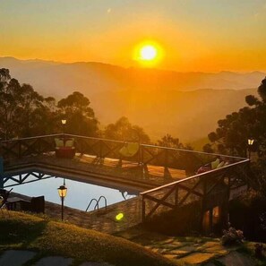 A sunset view from a bridge overlooking a valley with a pool and a gazebo. - Pousada Village Pôr do Sol (Campos do Jordão)