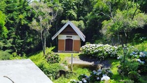Uma pequena estrutura de madeira com telhado inclinado, rodeada por vegetação exuberante e plantas floridas.