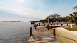 Un muelle de madera se extiende sobre aguas tranquilas, con una playa de arena y palmeras a la derecha.