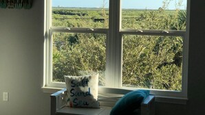A room with a large window overlooking a field. A pillow with the text "Salt Sand Sea" is placed on the windowsill.