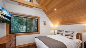 Ein Schlafzimmer mit großem Fenster, Holzbalkendecke, einem Bett mit weißer und gestreifter Bettwäsche und einem Nachttisch mit Lampe.