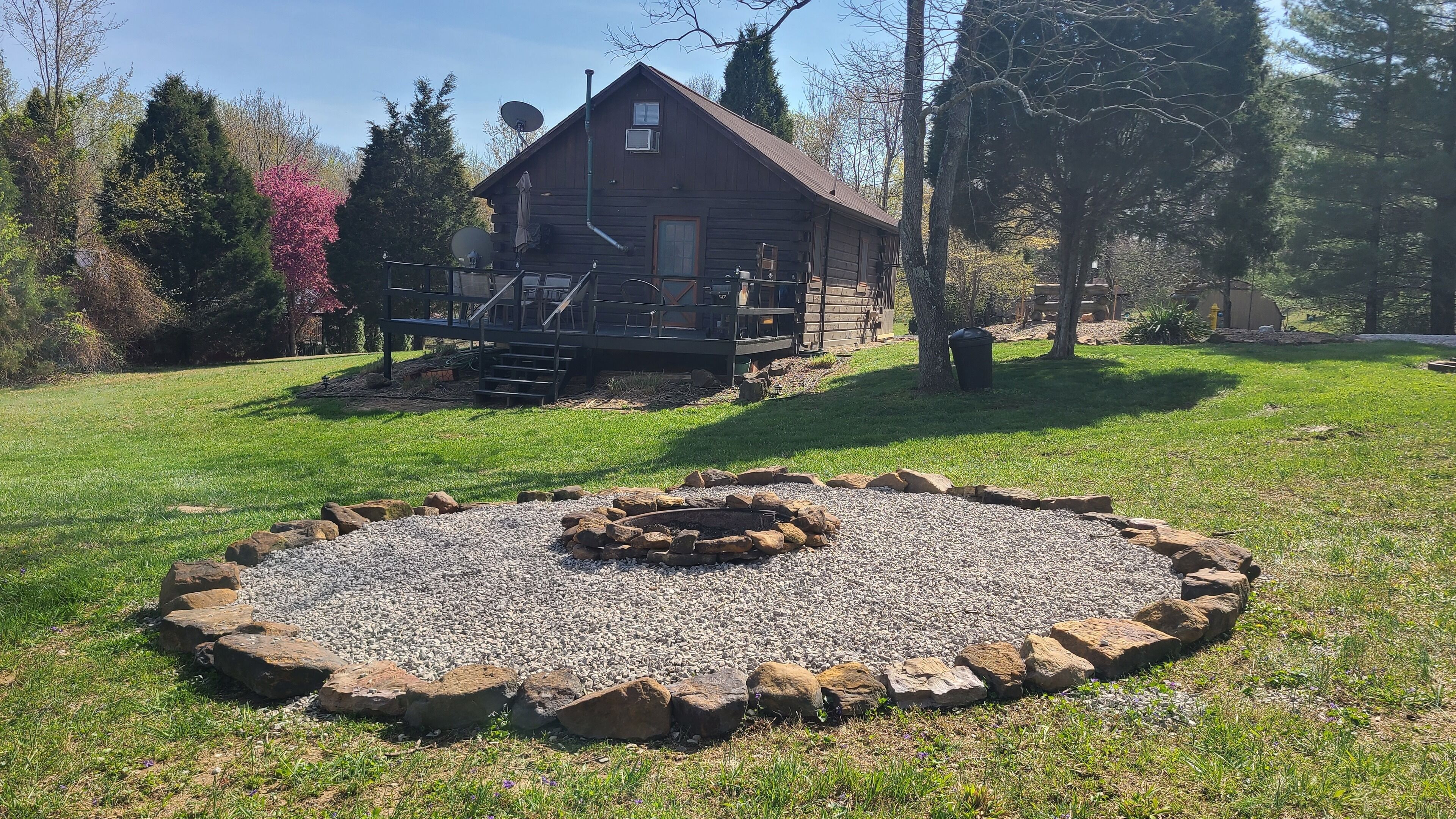 A rustic cabin with a deck, surrounded by a well-maintained lawn and a circular stone fire pit.