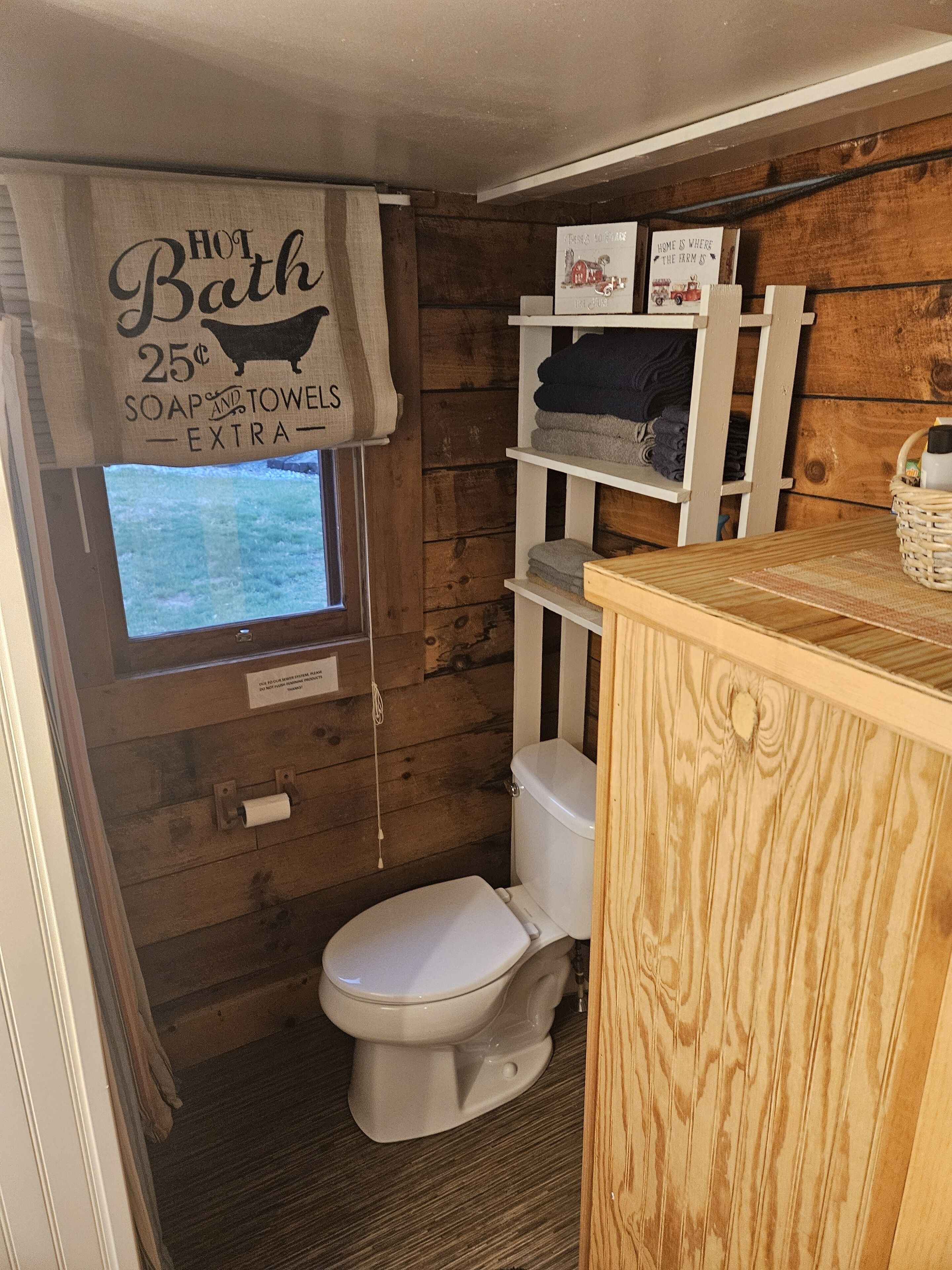 Ein kleines, rustikales Badezimmer mit Holzvertäfelung, einer Toilette, Regalen mit Handtüchern und einem Schild, das zusätzliche Seife und Handtücher für 25 Cent anbietet.