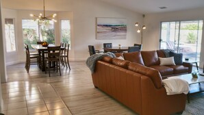 A spacious living area with a brown leather sofa, dining table with chairs, and a large window providing a view of the outdoors. - Posh Villa Sleeps 12 w/ 4 bedroom, 3 bath Pool & Spa (Indio)