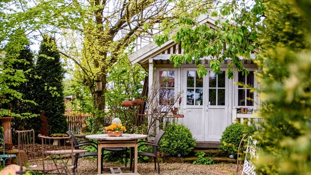 A rustic garden with a wooden table and chairs, surrounded by greenery and a white building with windows. - Werts HOF Ferienwohnung 2 (Rauschenberg)