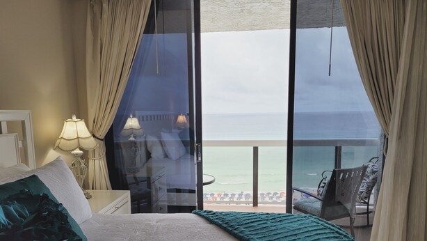 A bedroom with a large bed, a nightstand, a lamp, and a view of the ocean through a glass door. - Coastal Bliss and Million Dollar Views (Miramar Beach)