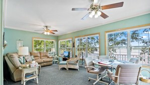 A spacious living room with a large window, a ceiling fan, a sofa, a coffee table, and a desk with a chair. - Lakefront Cottage on Webster Lake with Beautiful Sunsets, 2 bed, 1 bath (North Webster)