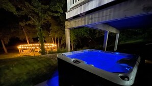 A hot tub with a wooden deck and a bench, surrounded by a well-lit patio area with string lights.
