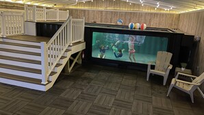 An indoor swimming pool area with a wooden floor, a poolside seating area, and a large screen displaying underwater scenes.