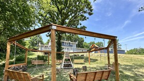A wooden swing set with benches and a ladder in a grassy area with a house in the background.