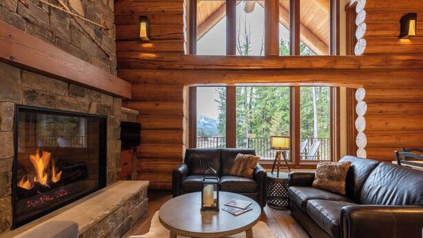 L’intérieur chaleureux d’un chalet en bois, avec une cheminée en pierre, des canapés en cuir et de grandes fenêtres donnant sur le paysage extérieur.