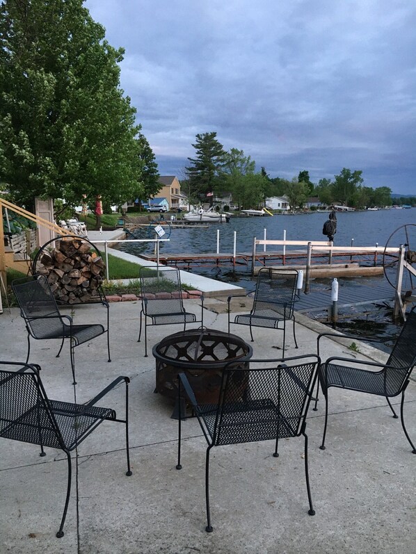 An outdoor patio with metal chairs and a fire pit, overlooking a body of water with boats and houses in the background.
