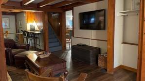 Ein gemütliches Wohnzimmer mit Holzboden, einem an der Wand befestigten Fernseher, einem Essbereich mit Stühlen und Tisch sowie einer Treppe zu einer weiteren Ebene.