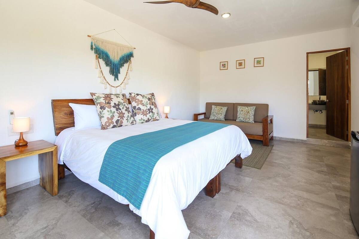 A bedroom with a bed, bedside tables, a sofa, a hanging macrame wall decoration, and framed pictures on the wall.