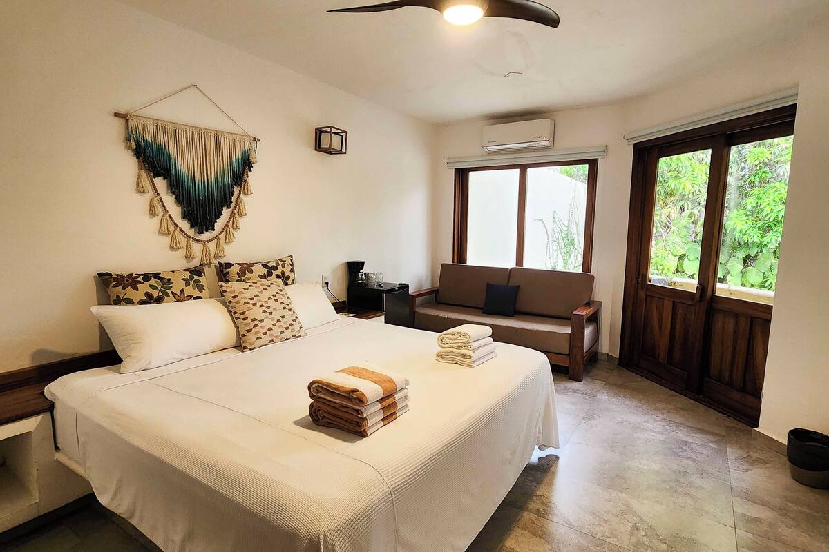 A hotel room with a large bed, a sofa, a small table, and a window with a view of greenery.
