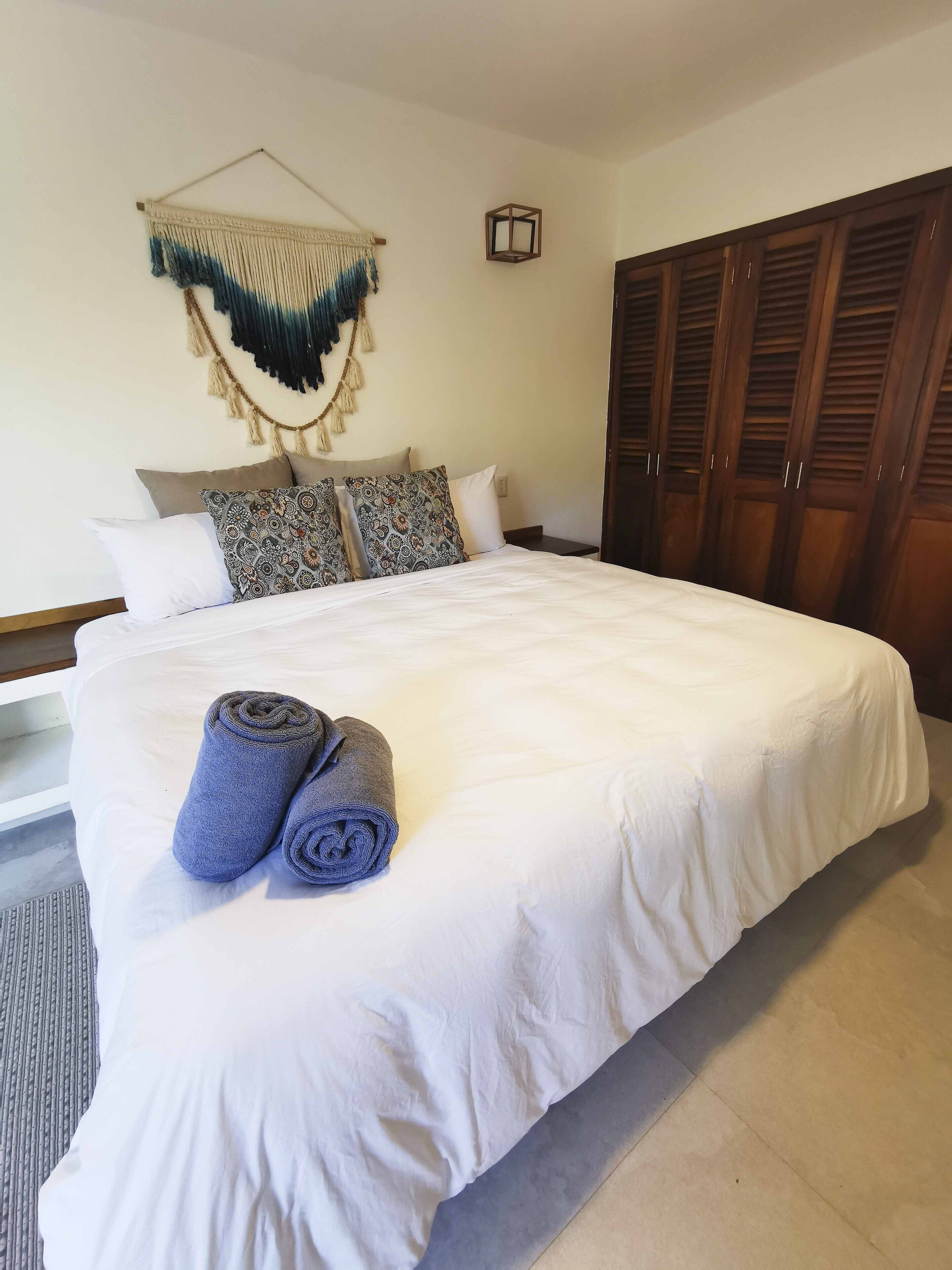 A neatly made bed with white linens and two rolled towels, a wall-mounted macrame piece, and a wooden wardrobe.