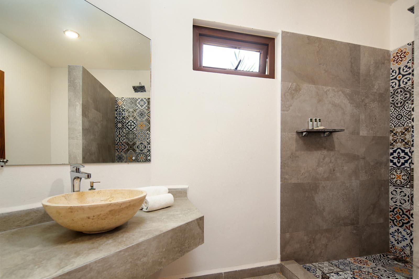 A bathroom with a large bowl sink, a mirror, a shower with patterned tiles, and a small window.