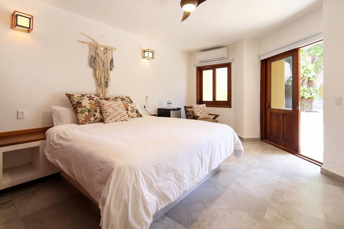 A bedroom with a large bed, a bedside table, a chair, a small table, and a window with a view of greenery.