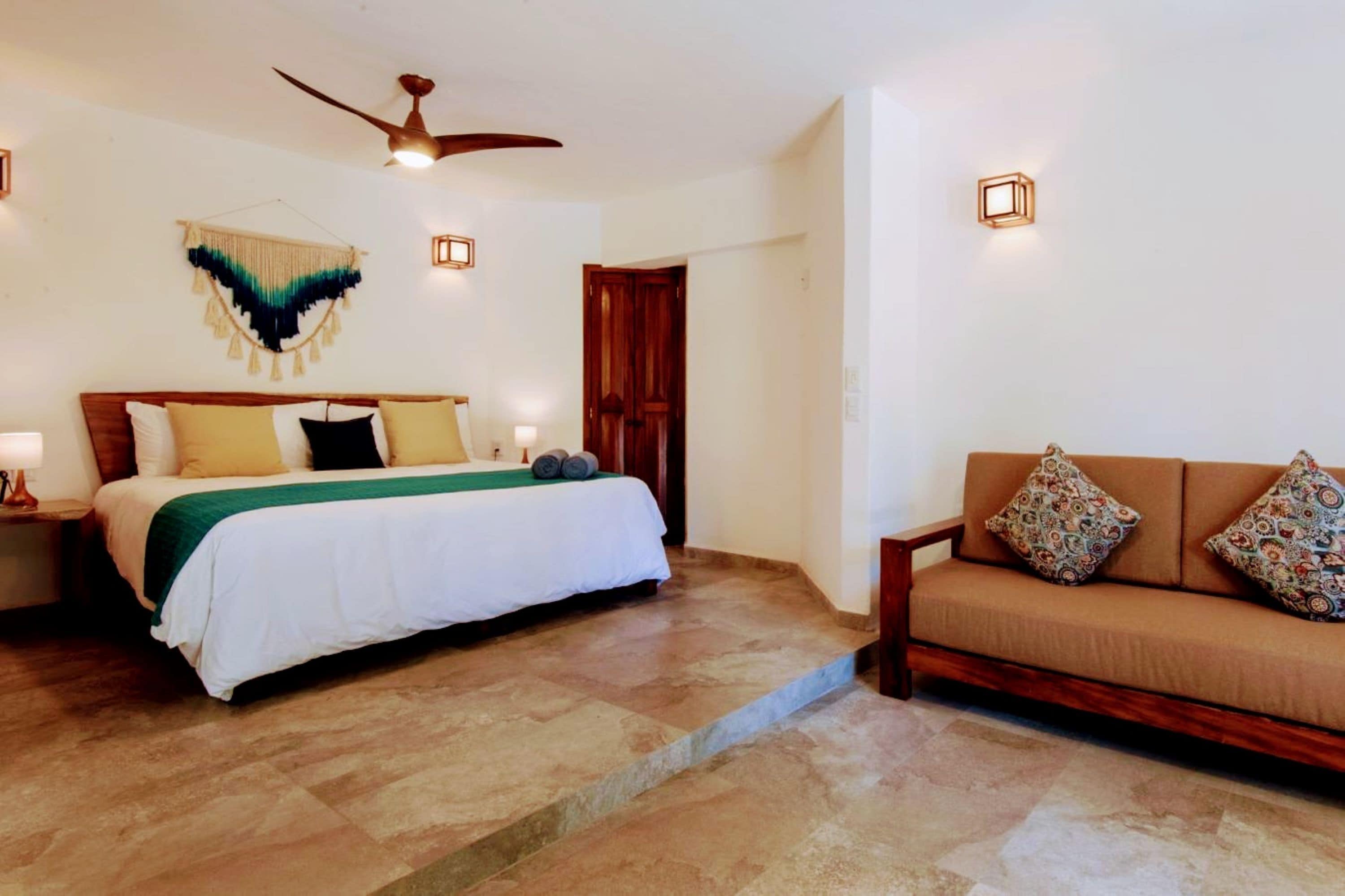 A bedroom with a bed, a wooden headboard, a green throw blanket, a wooden bench, and a wall-mounted light fixture.