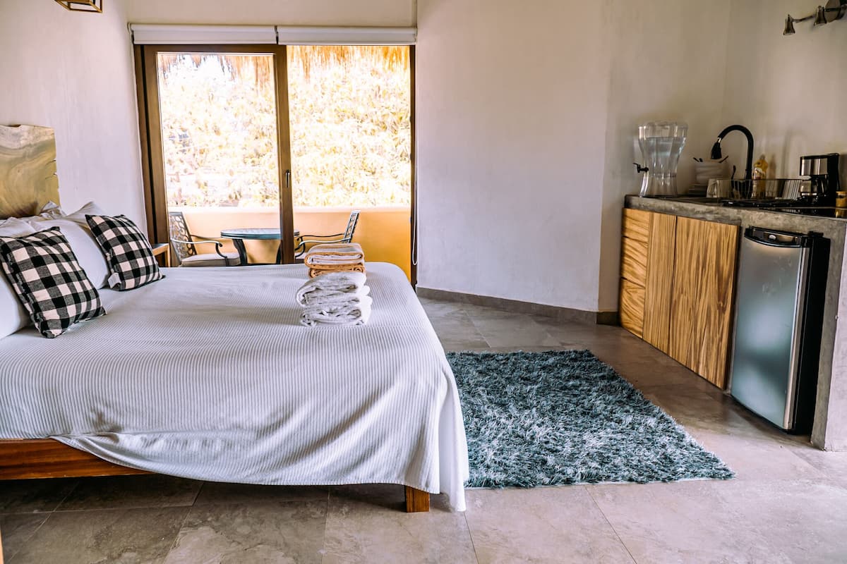 A bedroom with a bed, a small table, a chair, a kitchen area with a refrigerator, and a view of trees through a sliding door.