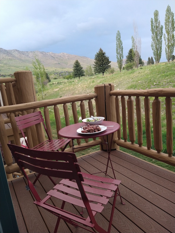 A wooden deck with a table and two chairs, overlooking a grassy landscape with trees and hills. - Powder Mountain Family Friendly Condo @ Moose Hollow Sleeps 6! (Eden)