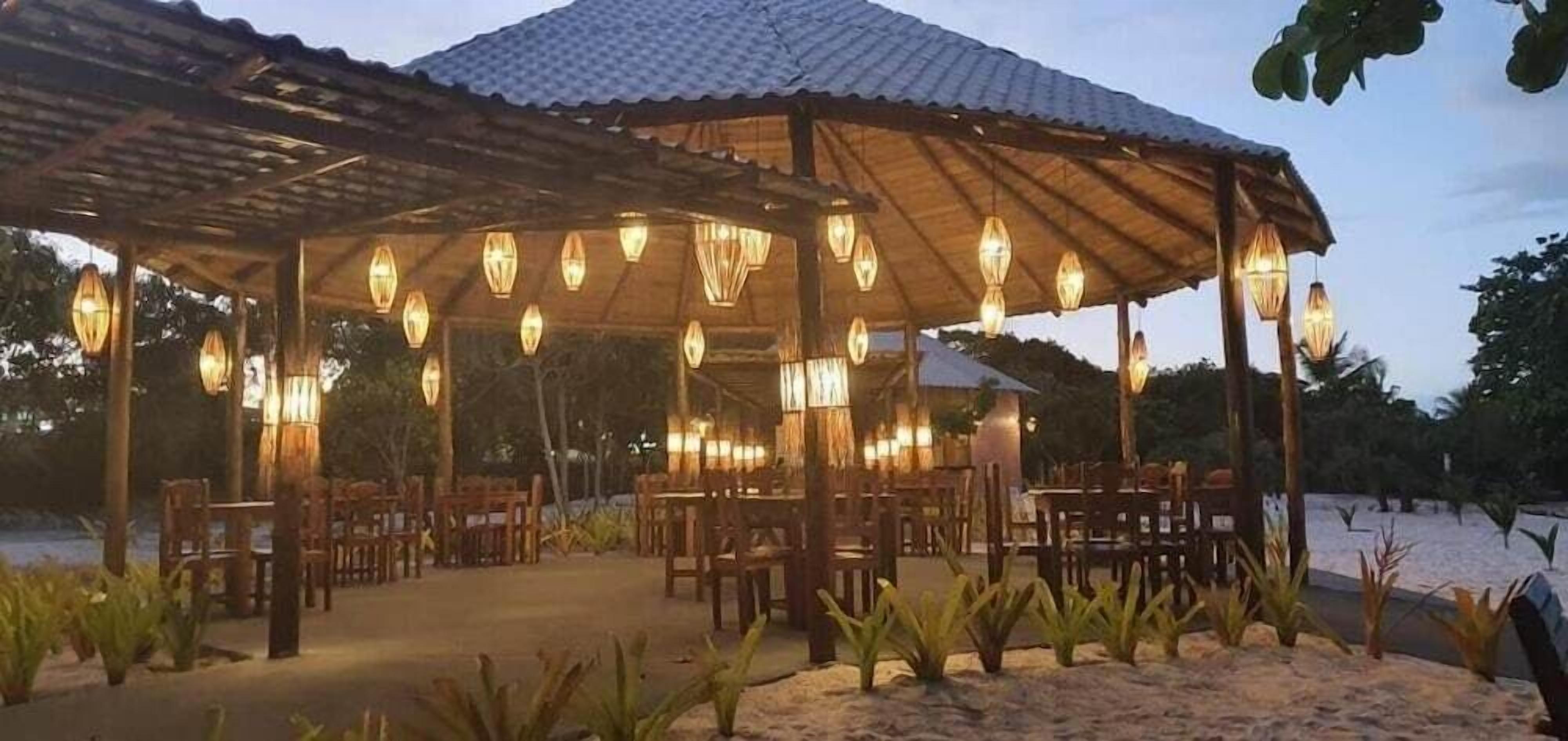 Uma área de refeições ao ar livre com um pavilhão coberto, várias mesas e luminárias pendentes em estilo lanterna.