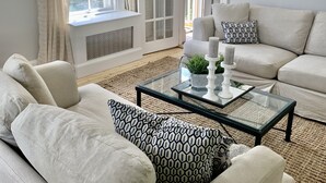 A living room with a beige sofa, patterned cushions, a glass coffee table, and a view of the outdoors through a large window.