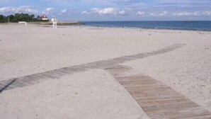 A beach with a wooden boardwalk leading to the water, a lifeguard tower, and a few buildings in the background.