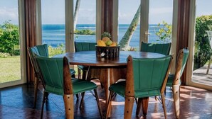 A round wooden table with green chairs, a fruit bowl, and a view of the ocean through large windows. - Malaqereqere Ocean View, Villa 2 (Sigatoka)