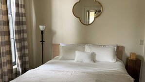A neatly made bed with white linens, a round mirror on the wall, and a floor lamp beside the window.