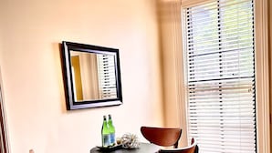 A dining area with a round table, four chairs, a framed mirror, and a window with blinds.