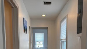 A narrow hallway with wooden flooring, white walls, and a door leading to a bedroom with a bed and window blinds.
