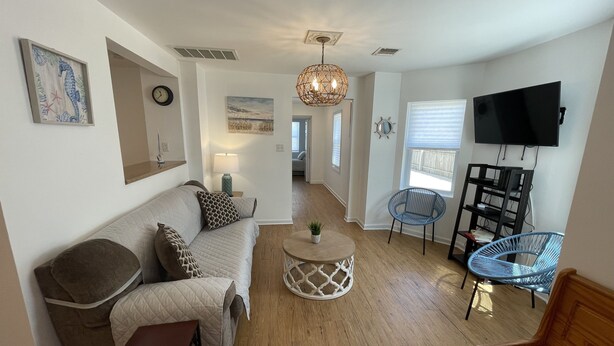 A modern living room with a sofa, coffee table, and chairs. There is a TV mounted on the wall and a window with blinds.