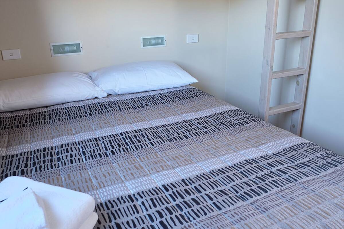 Un lit simple avec une couvre-lit à motifs, un oreiller blanc et une échelle en bois appuyée contre le mur.