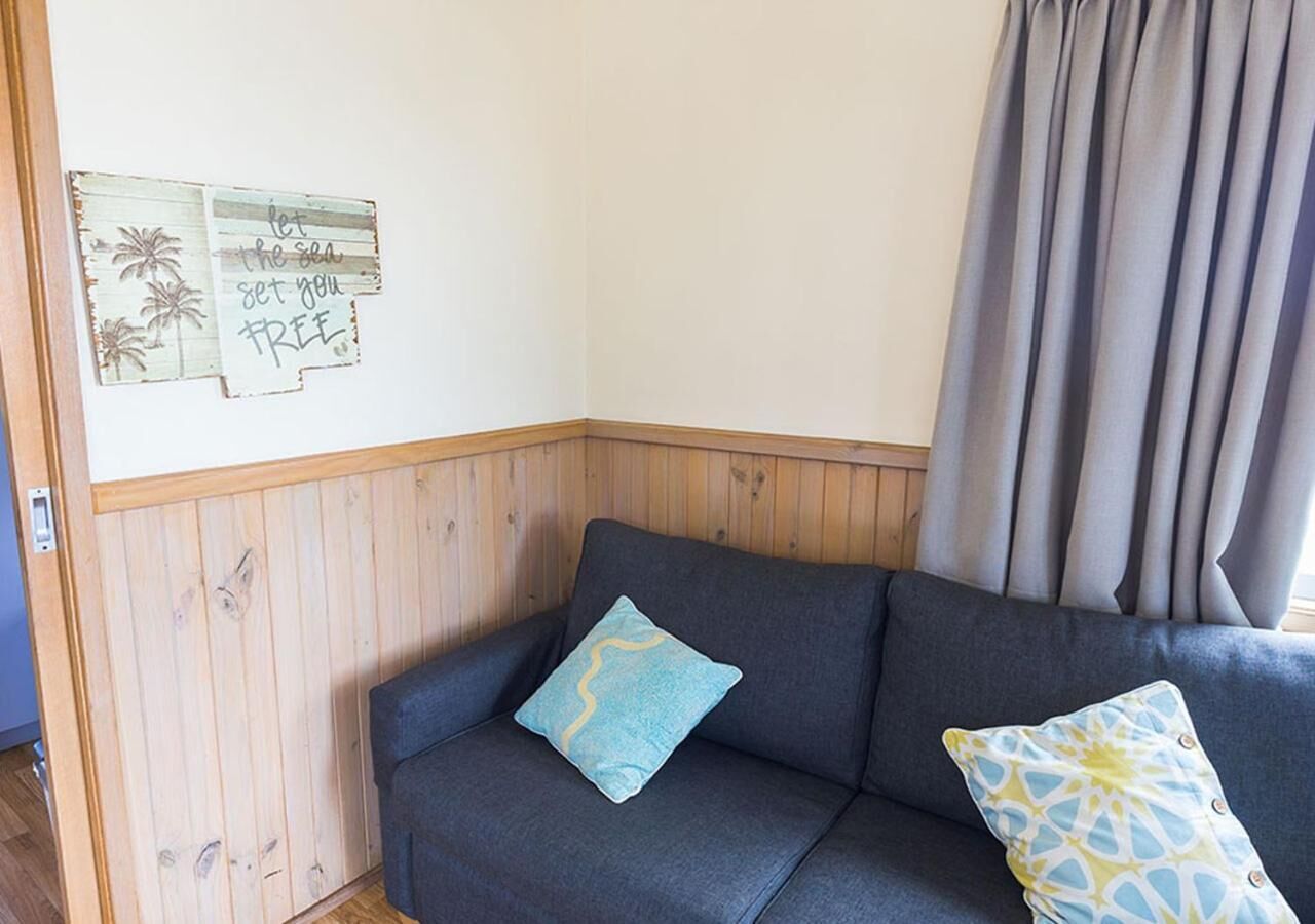 Ein Eckzimmer mit einem dunkelgrauen Sofa, zwei gemusterten Kissen, Holzvertäfelung und einer Wand mit tropischem Kunstwerk.
