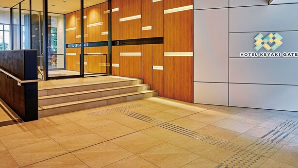 A modern hotel lobby with a wooden reception desk, a staircase leading to the upper floor, and a sign reading "HOTEL KEYAKI GATE".