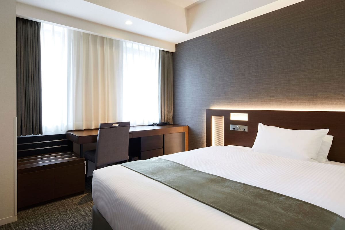 A hotel room with a bed, desk, chair, and a view of the outdoors through the window.