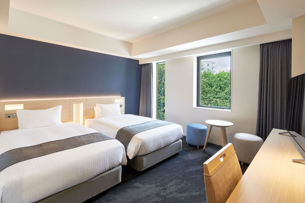 A hotel room with two beds, a small round table, a desk, and a window with a view of greenery.