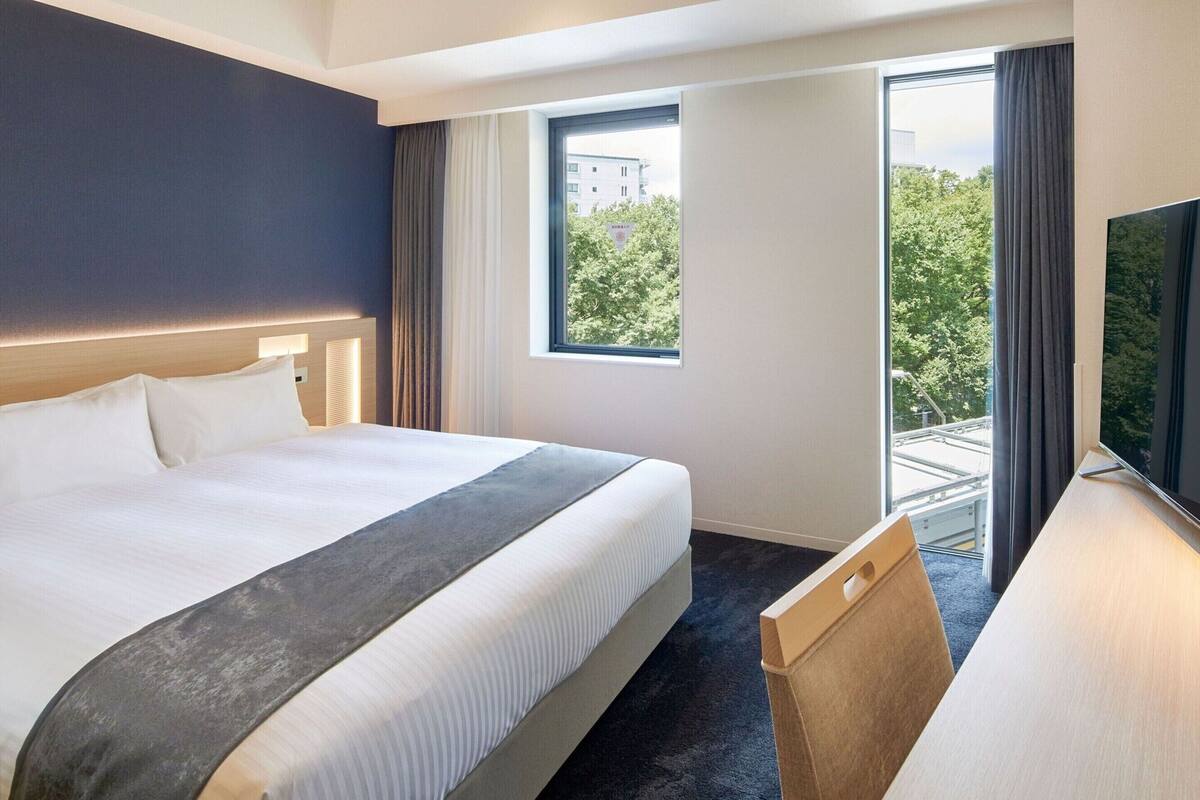 A hotel room with a large bed, a desk, a TV, and a window with a view of trees and buildings.