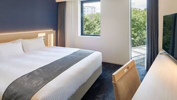 A hotel room with a large bed, a desk, a TV, and a window with a view of trees and buildings.