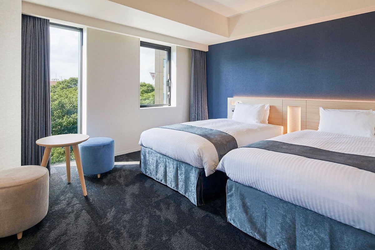 A hotel room with two beds, a small round table, and a view of greenery outside.