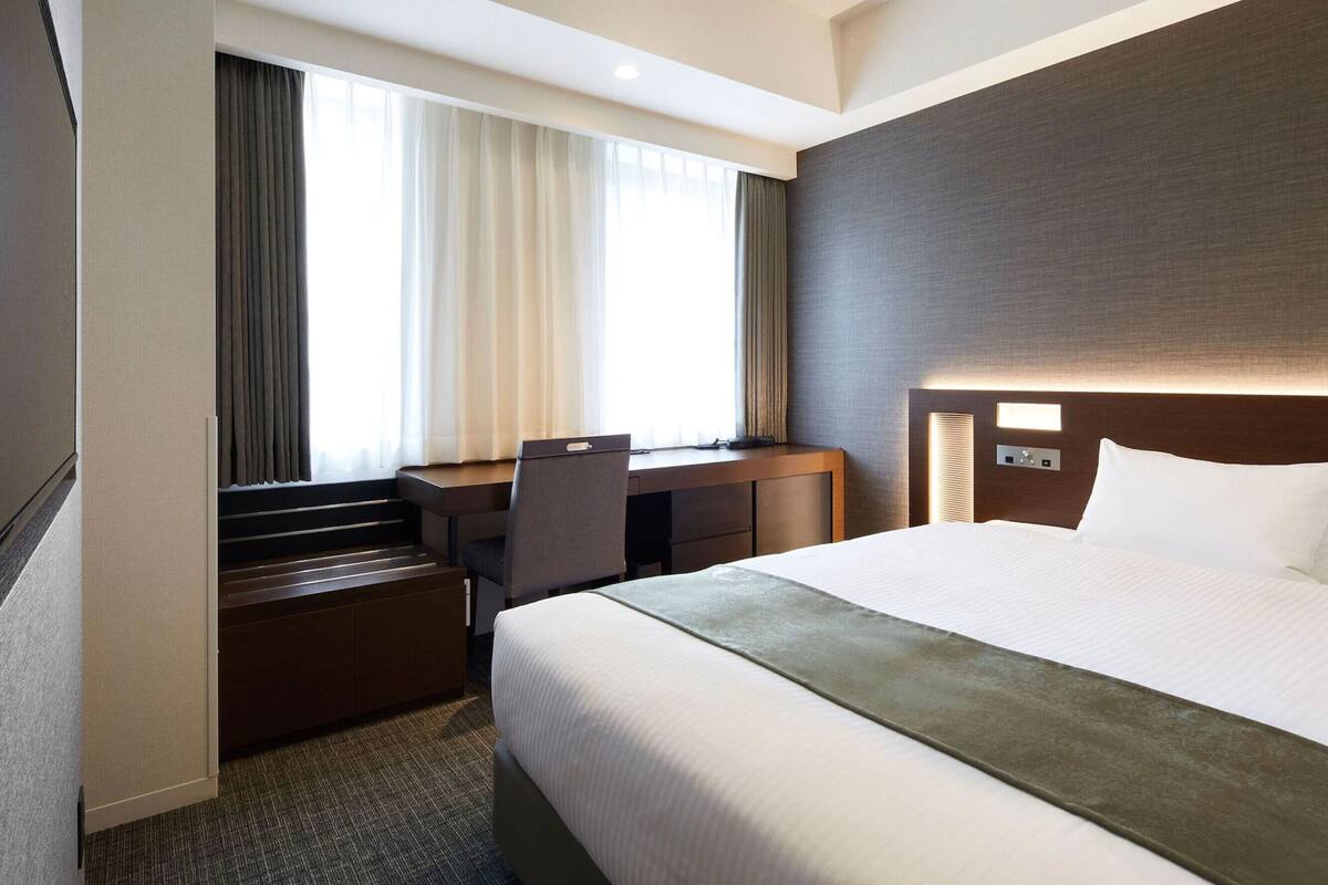 A hotel room with a bed, desk, chair, and a view of the outdoors through the window.