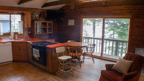 Ein Ferienhaus-Innenraum mit Holzausbau, einer Küchenzeile, einem Esstisch mit Stühlen und einem großen Fenster mit Blick auf eine Terrasse.