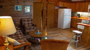 Ein gemütliches Blockhaus-Interieur mit einem Wohnbereich, der ein Karomuster-Sofa, einen runden Couchtisch und eine Lampe beherbergt. Ein Küchenbereich mit Holzschränken und einem Kühlschrank ist ebenfalls zu sehen.