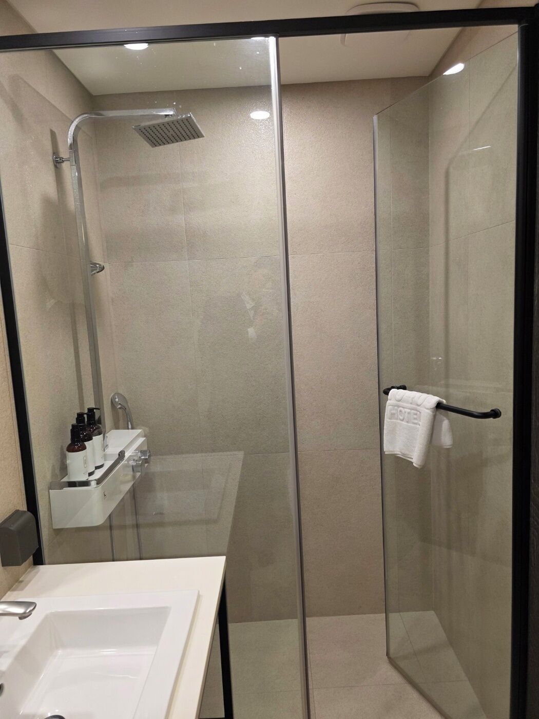 Ducha moderna con mampara de vidrio, un lavamanos blanco y un perchero con toalla de hotel.