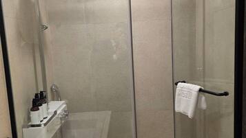 Ducha moderna con mampara de vidrio, un lavamanos blanco y un perchero con toalla de hotel.