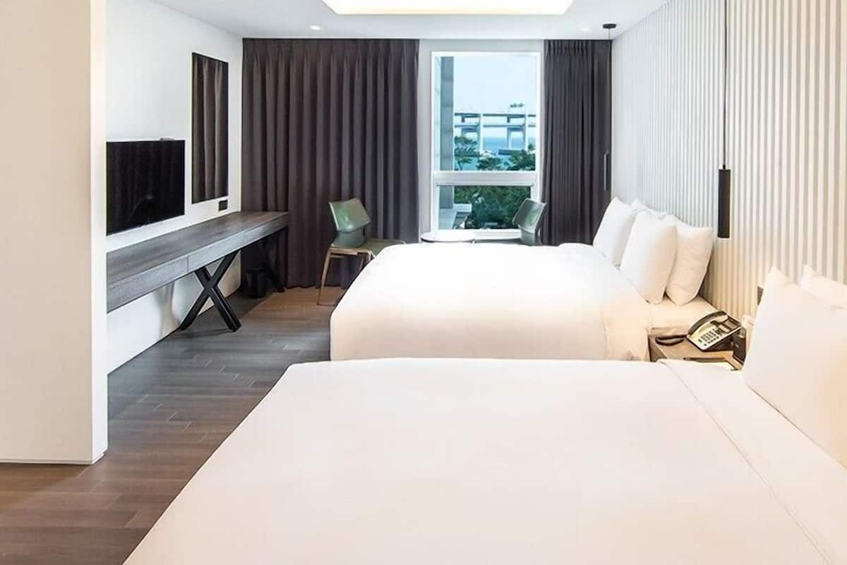 A hotel room with two beds, a desk with a chair, a TV, and a large window with a view.