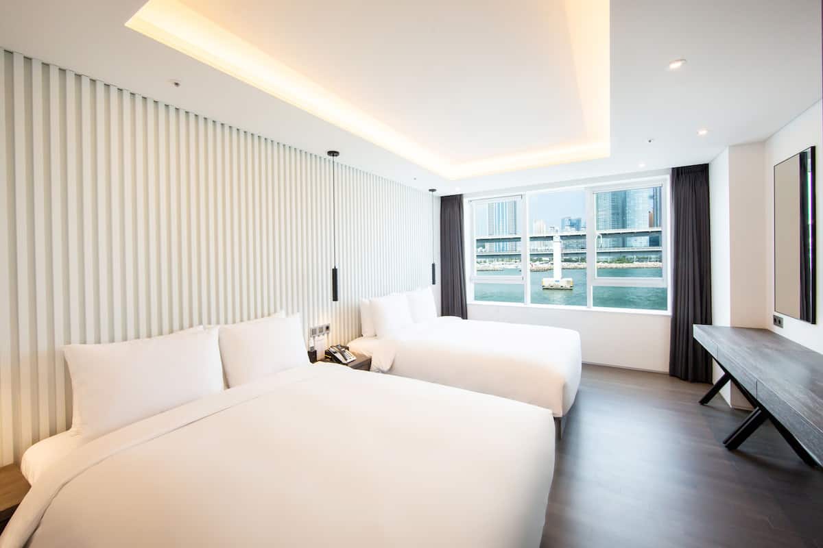A modern hotel room with two beds, a large window with a city view, and a flat-screen TV.