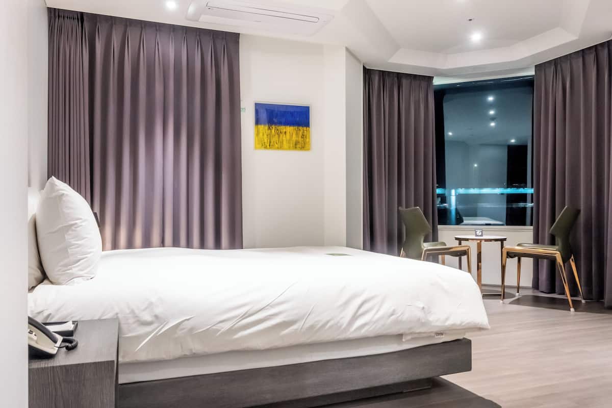 A modern hotel room with a large bed, grey curtains, a small table with chairs, and a window with a view of the cityscape.