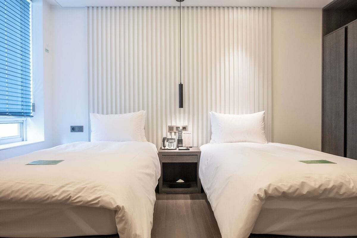 A hotel room with two beds, a bedside table with a phone and a lamp, and a window with blinds.